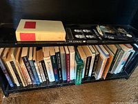 Photo of lower bookshelf filled with a collection of approximately 35-40 books, mostly paperbacks with some hardcover books, showing titles related to biography, law, and other non-fiction genres.