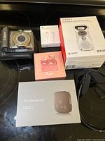 Photo showing Canon PowerShot A590 digital camera, Apple Lightning to USB cable box, iLuv wireless earbuds box, Eton hand turbine radio box, and TV Connector box on black surface.