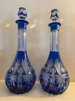 Front view of two cobalt blue cut to clear crystal decanters with clear geometric stoppers showcasing floral and foliage etching patterns on the bulbous bases.