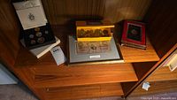Overview of coin and currency collection displayed on wooden shelf with various items including framed currency, coin sets in boxes, and loose coins in packaging.