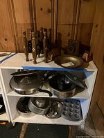 Full view of shelving unit displaying collection: wooden stand, copper cowbells, brass dish, metal bookend, assorted pots, muffin tin, pie pans, and lids.