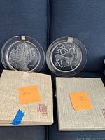Three clear crystal Lalique Paris plates featuring detailed etched animal designs from 1968 and two from 1972 alongside their original textured beige boxes labeled with the dates.