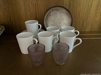 Front view of six white porcelain coffee/tea cups, two frosted purple glass cups, and one metal plate arranged on a wooden shelf.