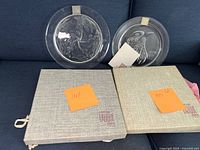 Two Lalique crystal plates displayed on couch with original presentation boxes beneath, showing labels with production years 1967 and 1973.