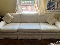 Front view of the light beige upholstered sofa showing the three seat cushions, subtle leaf pattern fabric, and two matching square pillows.