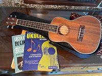Single photo showing the whole Lohanu ukulele lying on its black soft case with two beginner ukulele books laid out in front of it.