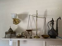 Shelf display showing various brass and mixed metal Middle Eastern decorative items including the large ewer, balance scale, smaller vessels and figurines arranged together.
