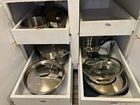 Two open drawers showing multiple stainless steel pots and pans, some stacked, several lids on top or in bottom drawer.