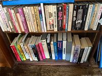 Photo of two shelves filled with approximately 40-50 books of mixed genres and formats including paperbacks and hardcovers, showing spines with various titles and authors.