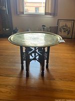 Full view of the brass tray table on folding wooden stand placed on hardwood floor with various household items in background.