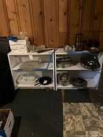 Pair of white wooden bookshelves placed side by side with various kitchen and household items around and on them.