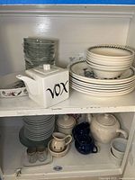 Photo showing mixed dinnerware on a white shelf. Includes stacked floral pattern ceramic plates and bowls with green leaf borders (Portmeirion Botanic Garden), clear glass bowls, a white ceramic teapot with black lettering, small dark blue mugs, and other rustic style ceramic items (Hartstone).