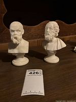 Two white plaster busts of ancient Greek figures Socrates (left) and Homer (right) on round pedestal bases, about 6 inches tall, displayed on a wooden surface with dark carved backrest.