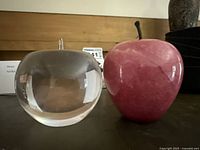 Front and side views of the clear Tiffany & Co. crystal apple and the pink Eikoh stone apple with dark brown stem on a dark surface in front of a wood backdrop.