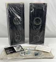 Two black rectangular speakers wrapped in plastic with accessories laid out below.