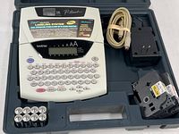 Brother P-Touch label maker with keyboard, display, power adapter, connection cable, label cartridge, and batteries arranged in original plastic storage case.