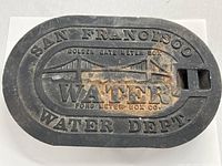 Top side of black oval cast iron water meter box cover showing embossed Golden Gate Bridge design, text SAN FRANCISCO WATER DEPT., GOLDEN GATE METER BOX, FORD METER BOX CO.