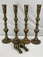 Four tall brass candle holders standing upright, showing turned design and bases.