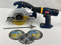 Front angle showing Ryobi cordless circular saw and drill with three circular saw blades and a saw guide on a white surface