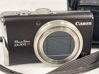 Front view of the Canon PowerShot SX200 IS camera with lens retracted showing branding and zoom specifications.