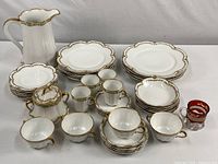 Full set of vintage white porcelain Theodore Havilland Limoges china with gold scalloped trim and floral vine pattern, cups and saucers arranged at front, plates stacked behind, water pitcher and covered sugar bowl included, cranberry souvenir cup at right front.