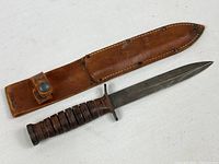 Full view of WWII military fighting knife alongside brown leather sheath on white background.