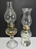 Two clear glass oil lamps with chimneys displayed side by side