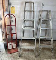 Front view of two aluminum step ladders beside red steel hand truck