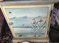 Front of cabinet showing hand-painted landscape, flowers, spider webs and insects