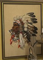 Framed needlepoint portrait of Indigenous chief, profile view with full headdress, gold frame visible