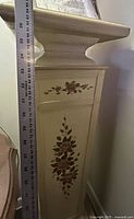 Front view of cream wood pedestal with floral painting and ruler showing height