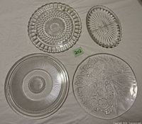 Four clear pressed glass serving pieces laid out: two round cake plates, one oval pickle plate, one round serving platter