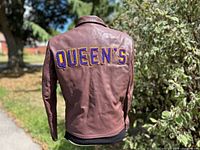 Back view showing QUEEN’S lettering on brown leather jacket