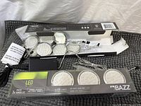 Retail box of three BAZZ LED puck lights and loose pucks with wiring and adapter