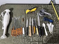 All items laid out: immersion blender, skewers, knives, sharpener, pizza cutter, can opener, measuring cups, spoons