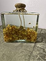 Full view of rectangular glass bottle with wooden cap, gold cord, and visible gold leaf in oil