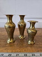 Three etched brass bud vases standing on wooden surface with ruler for scale