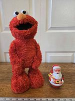 Tickle Me Elmo plush standing next to Santa roly-poly rattle, ruler for scale