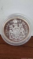 Reverse side of 1965 Canadian 50 cents coin showing Canadian coat of arms, date '1965', and 'CANADA 50 CENTS' inscription inside a clear round plastic case.