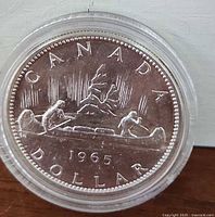 Reverse of 1965 Canadian silver dollar coin showing canoe with two figures and mountain scene, marking year 1965 and 'CANADA DOLLAR'.