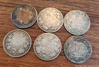 Six Canadian silver quarters placed on wooden surface showing reverse side with maple leaf wreath designs and dates including 1872. Coins show tarnish and wear.