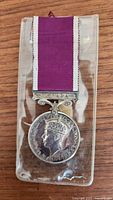 Overall view of medal with purple and white ribbon in plastic pouch