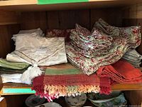 Overview of assorted linens on shelf: cream damask cloth, green napkins, red-ivory toile stack, solid red cloths, multicolour woven runner