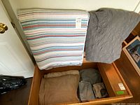 Striped quilt and grey quilted blanket draped over open trunk, additional folded linens inside