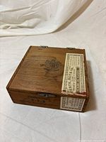 Angled view of wooden cigar box showing lid crest, label, and hinges