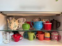 Entire lot on two cupboard shelves showing teapots, mugs, sugar shaker and Asian-style canister