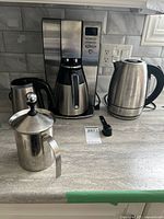 All items on countertop: Oster coffee maker, Chefman kettle, small insulated container, milk frother, scoop