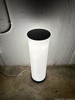 Full view of the cylindrical lamp illuminated, showing white frosted diffuser, black circular top and base, and cord on floor.