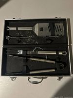 Photo of a silver metal carrying case containing three BBQ tools secured in black foam lining: a spatula, tongs, and a fork.