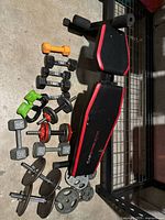 Overview of adjustable weight bench with CAP Strength branding and multiple dumbbells including neoprene-coated and metal dumbbells as well as various weight plates on the concrete floor.